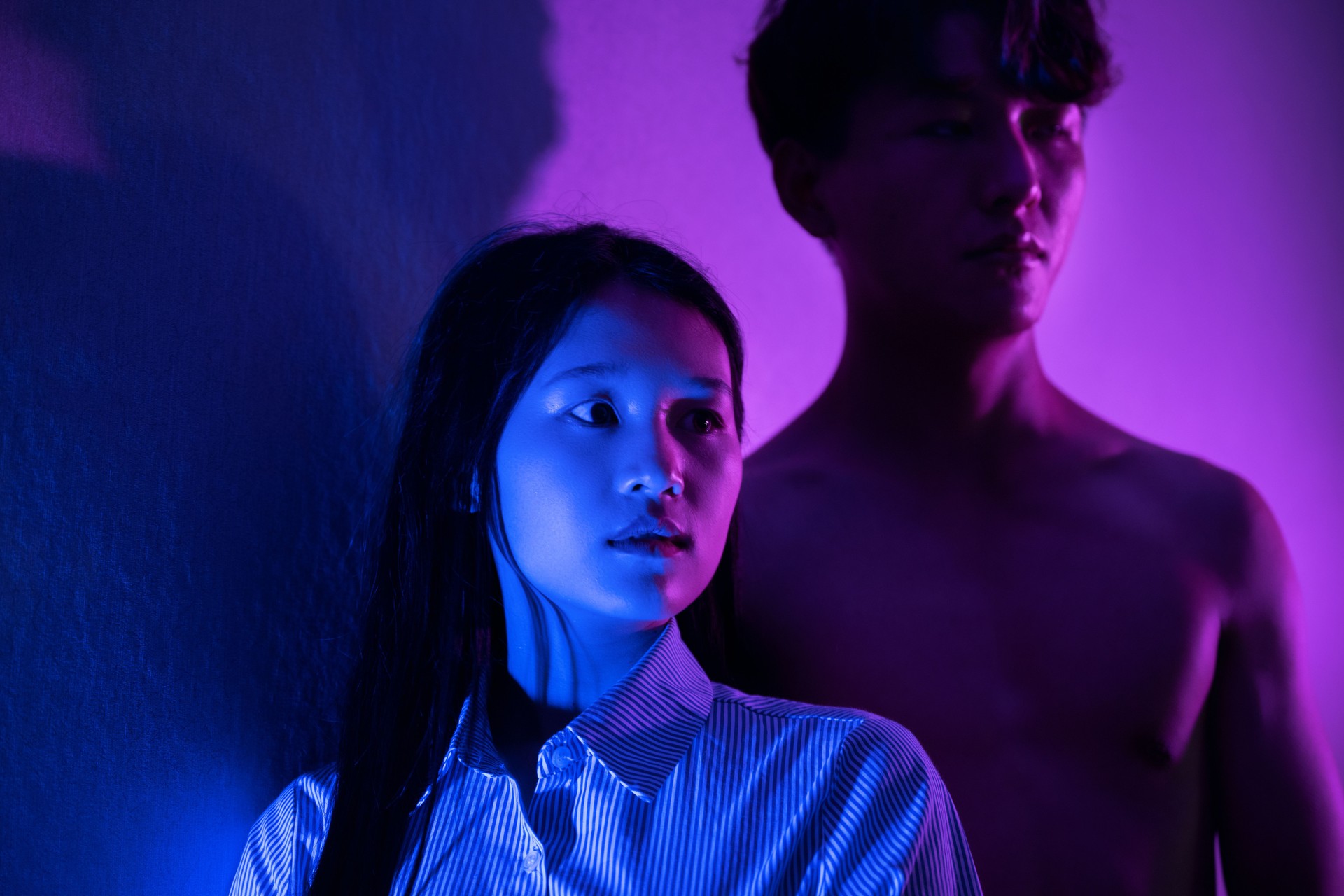 The bare muscles of a strong man in purple light stand next to a beautiful woman